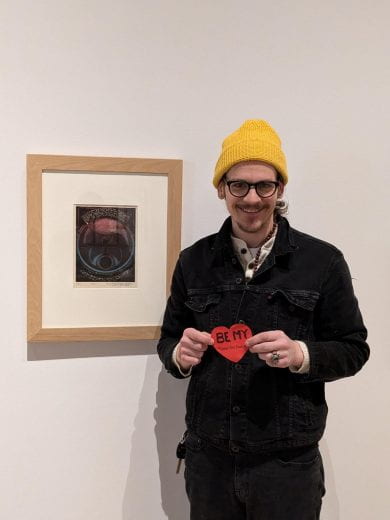 Beach SweetART contest participant holding a paper heart in front of a favorite artwork on display at the Beach Museum of Art.