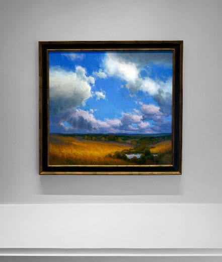 A fall landscape painting displayed on a wall with grass, bushes and shrubs along a creek, sky and clouds. 