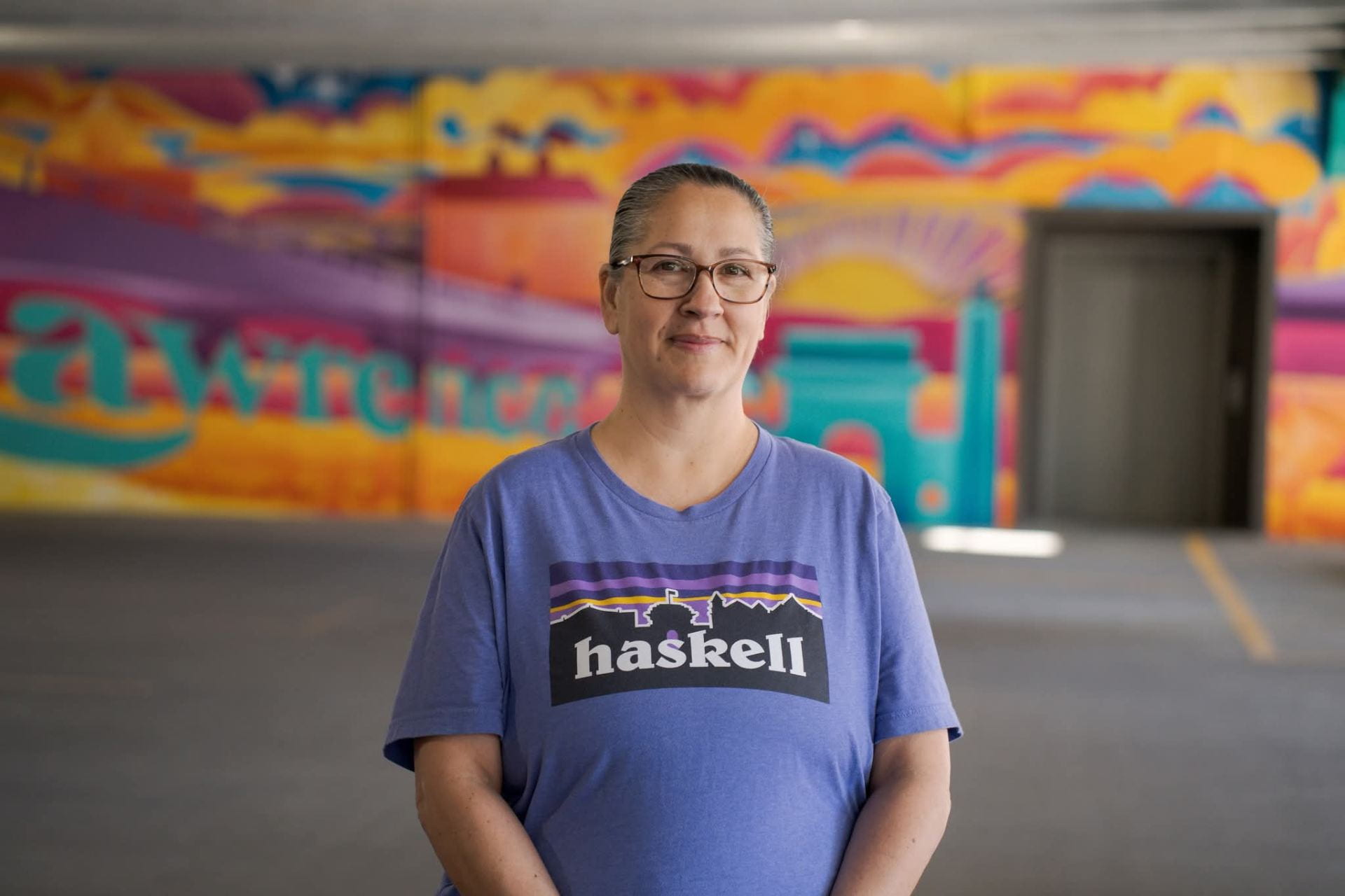 A woman standing in front of a painted mural. There is text printed on her t-shirt that reads "Haskell"