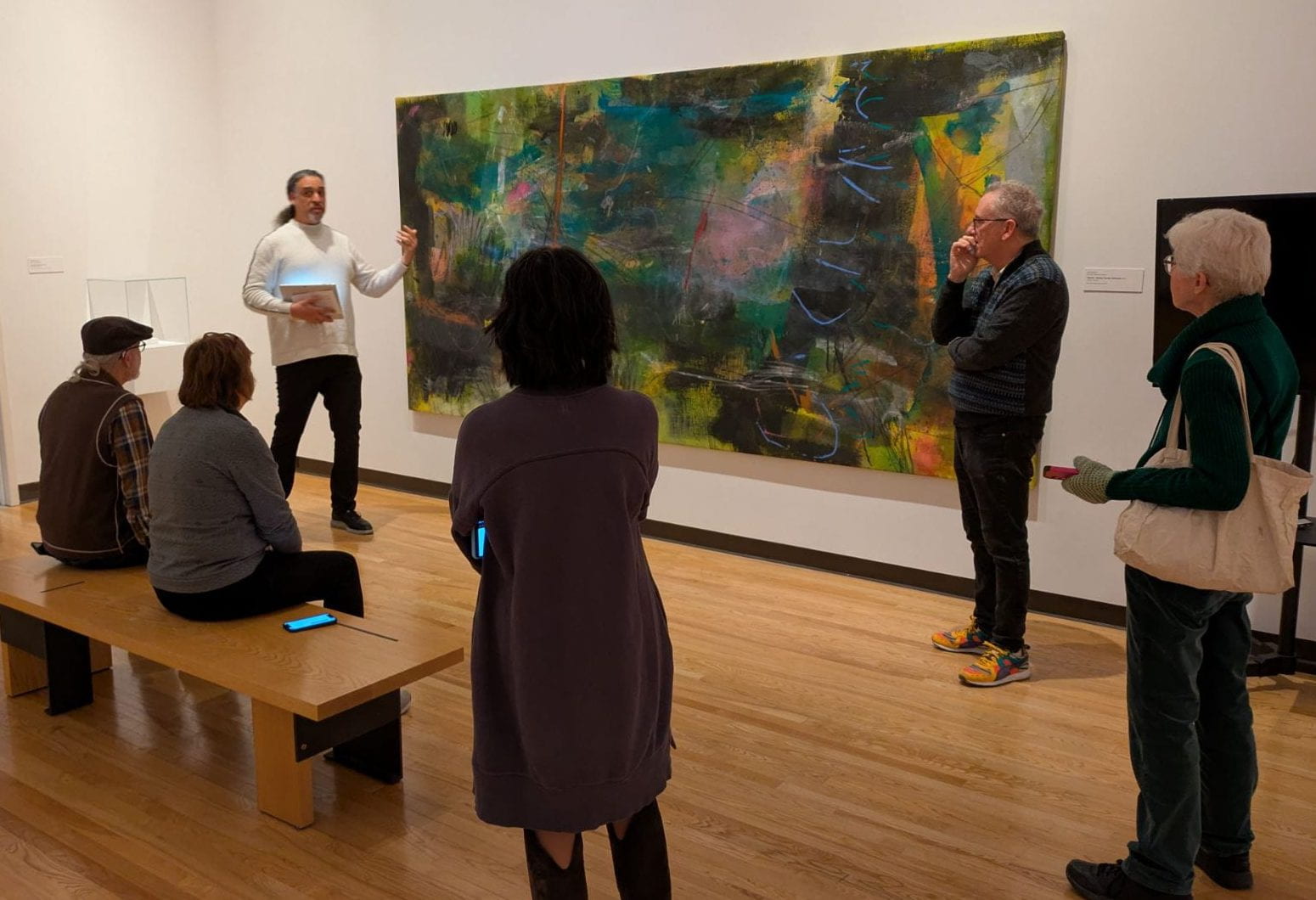 Participants gathered in a gallery and engaged in a discussion abotu an abstract colorful painting.