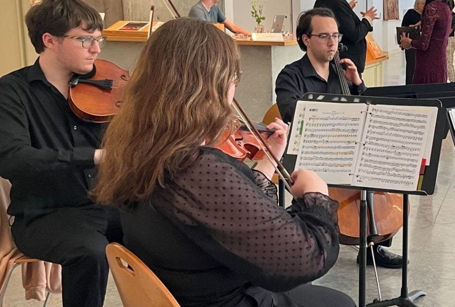 Students playing string instruments like violin and cello in the Beach Museum of Art atrium.