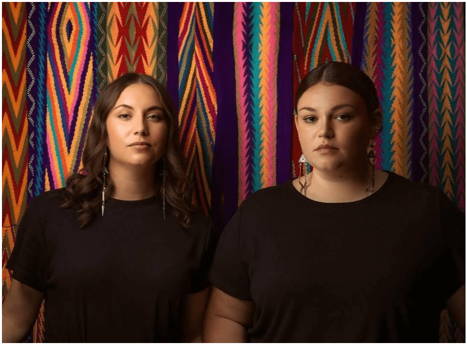 Two women looking in front. The background features various colorful belts from the Indigenous culture. Image credit: Ryan RedCorn, (Wahzhazhe/Osage Nation), "Courtney and Brittney Neff," 2021 dye-sublimation print on polyester fabric, 90 x 120 in. Image courtesy of and copyright by Ryan RedCorn