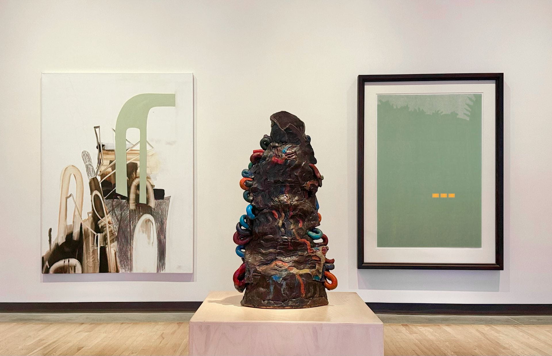 An exhibition gallery with two framed artworks displayed on a wall and an abstract sculpture resembling a tower displayed on a pedestal. 