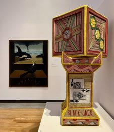 A view of the "STE[A]M" exhibitions at the Beach Museum of Art with a painting on a wall and a 3-D sculpture of a cube and pedestal made from corrugated cardboards with patterns on the surface.