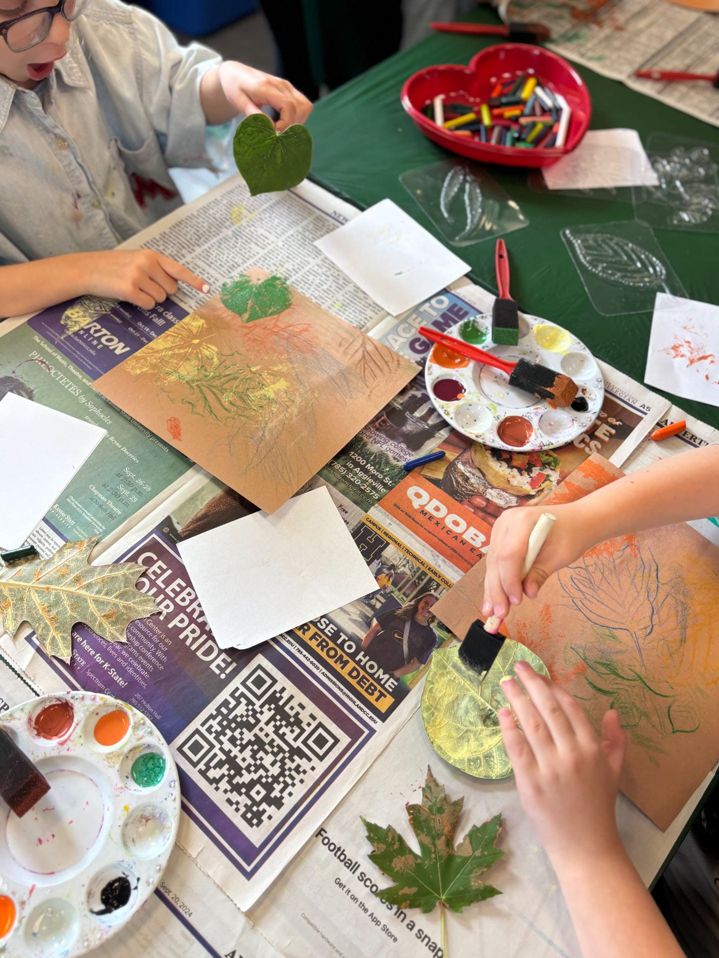 Children and painting on leaves to make leaf prints on paper. Paint brushes, paint and other art and craft materials are placed around. 