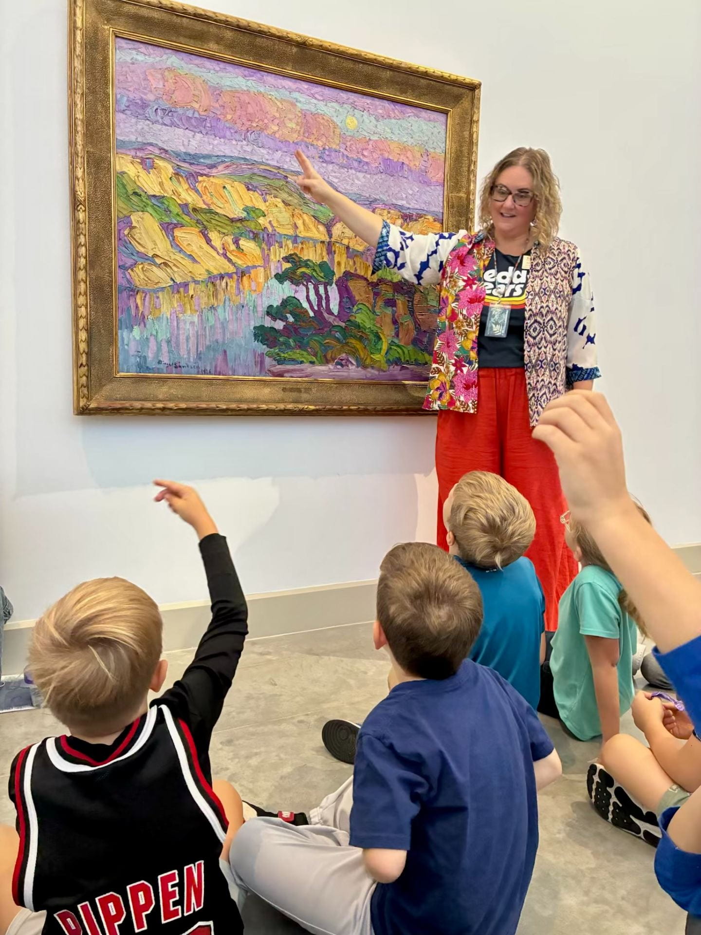 Art teacher and children discussing a painting on the wall. Teacher pointing at the painting and student raise their hands in response.