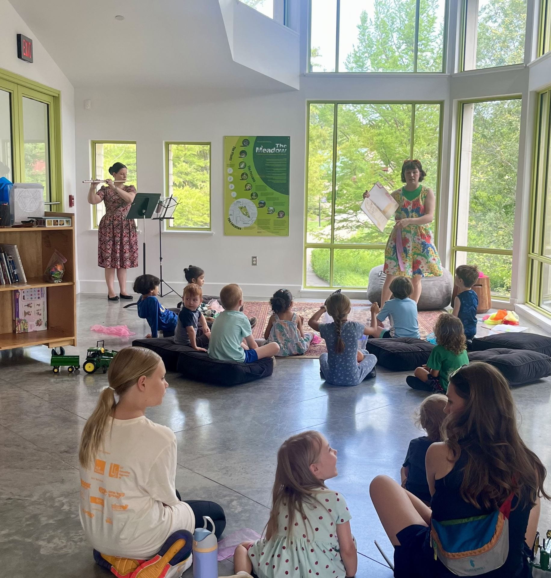 A woman reading a book to children and another woman playing flute.