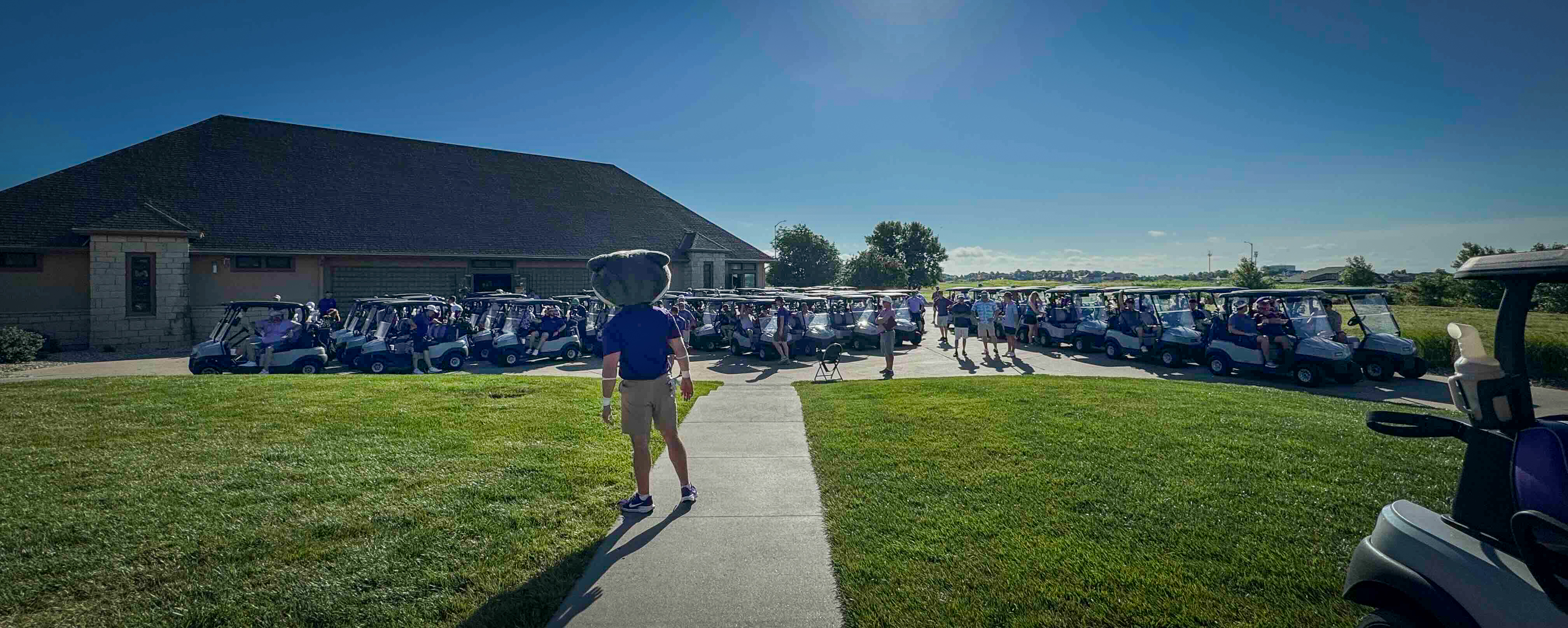 Willie the Wildcat, dressed in khaki shorts and a purple polo, stands in front of a group of purple golf carts with green grass and blue skies.