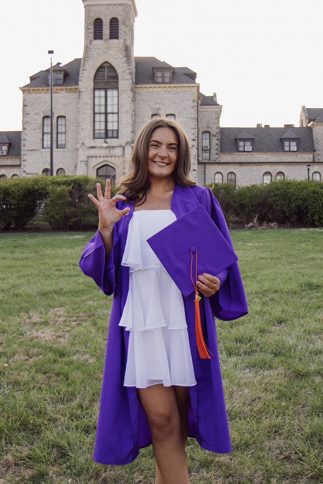 A young woman in a white dress and purple graduation gown standing in front of a camppus building and giving a Wildcat hand symbol.