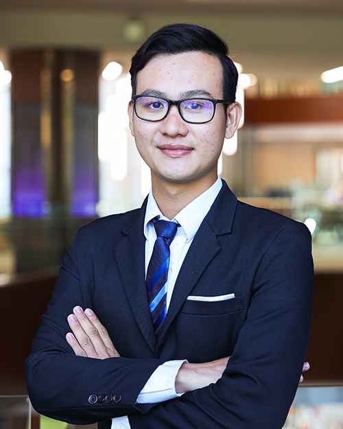 A student in a dark suit, blue tie and glasses crosses his arms and looks at the camera.