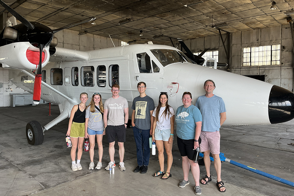 A professor and seve college students stand in front of a small, white plane in an airplane hanger.