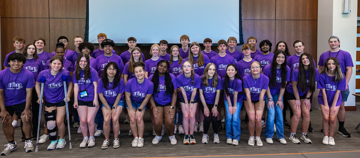 High school students wearing purple shirts pose for a group photo in a large conference room. 