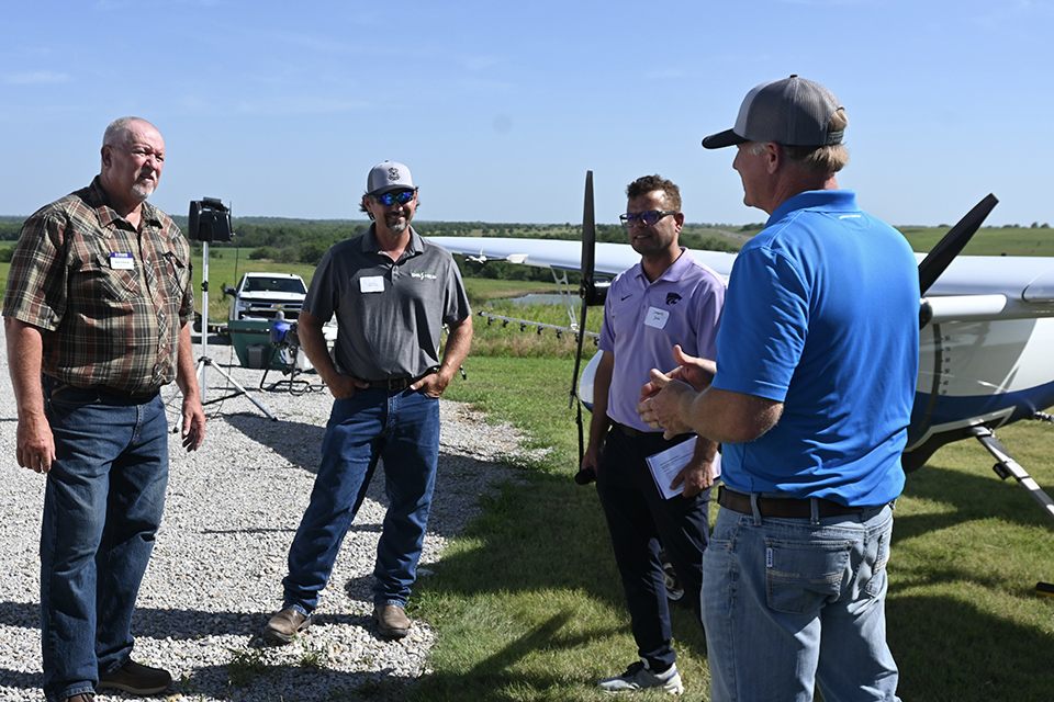 Four men in polo shirts and jeans stand around talking outside next to a crop plane. 