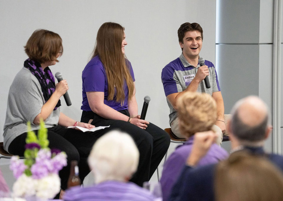 Three people sit at the front of a room talking on a panel. From left: A woman in a gray shirt and a black scarf with purple accents holds a microphone; a young woman in a purple shirt smiles and holds a microphone; and a young man in a gray and purple shirt speaks into a microphone while smiling.