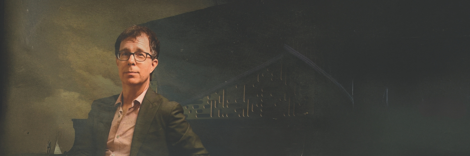 A man with dark hair and glasses sits on the left side of the frame wearing a blazer and light-colored shirt. Behind him and on the right side of the frame, dark colors and a triangular shape with circles and ovals in it.