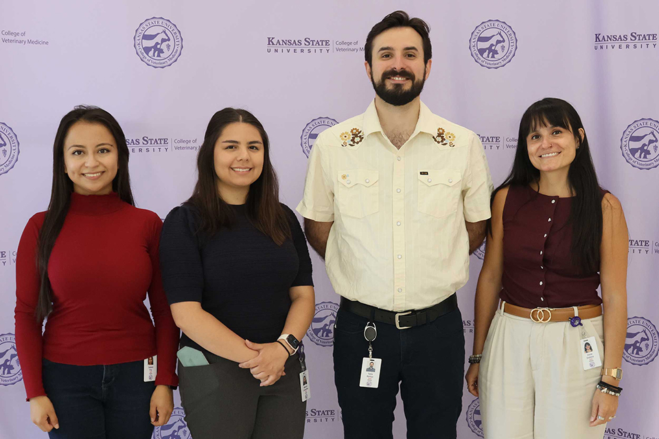 Four people stand in front of a Kansas State University College of Veterinary Medicine photo backdrop that is lavender with purple text and logos. 