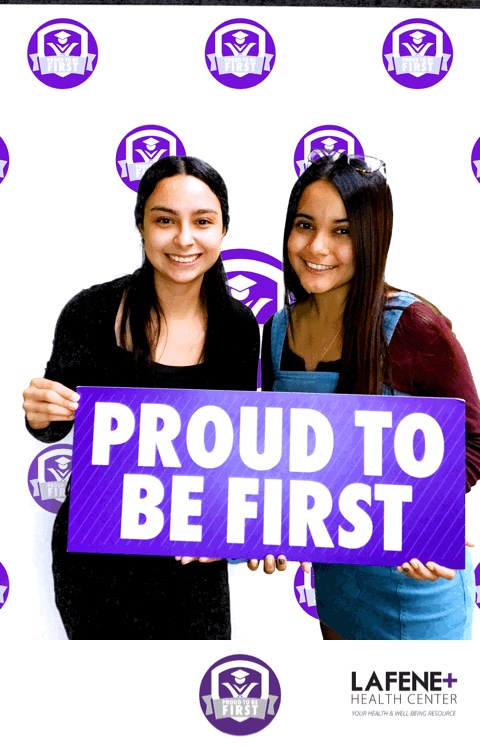 Two young women with dark hair smile at the camera and hold a sign that reads "PROUD TO BE FIRST" in front of a backdrop with the same message.