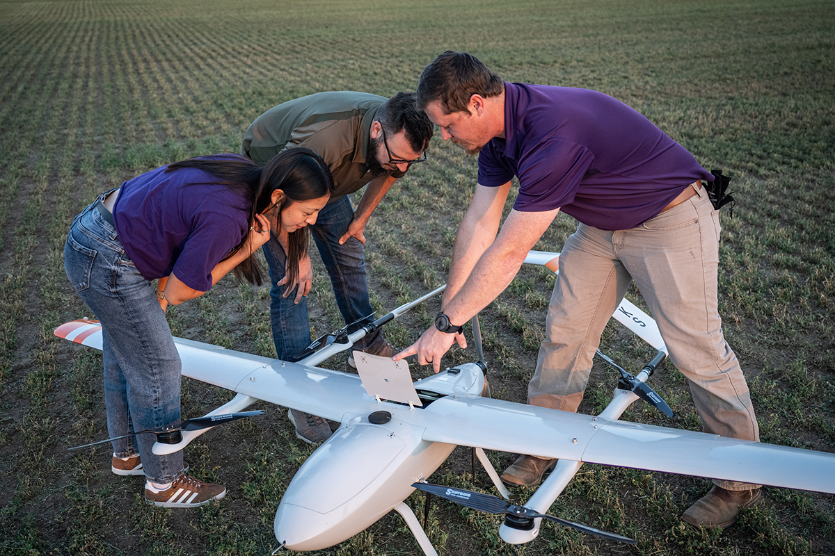 Three people, two men and one young woman, gather around an unmanned aircraft system that looks plane-like in a field. 