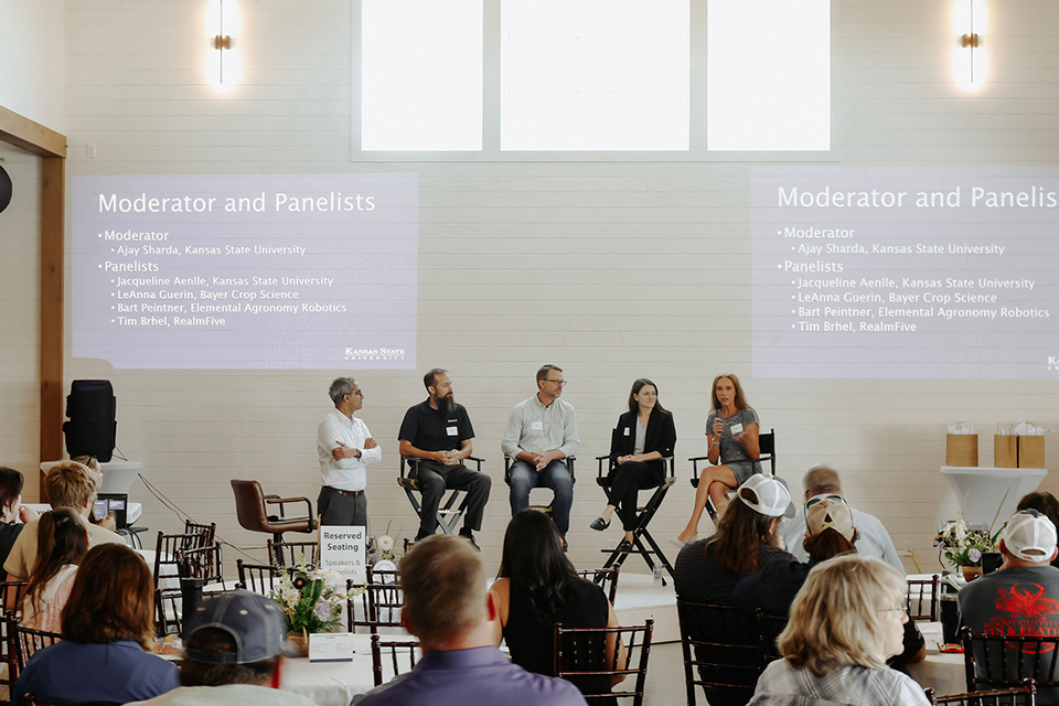 A photo shot from behind a crowd of people sitting and listening to a panel of speakers who are seated in director's chairs speaking in front of a white wall. 