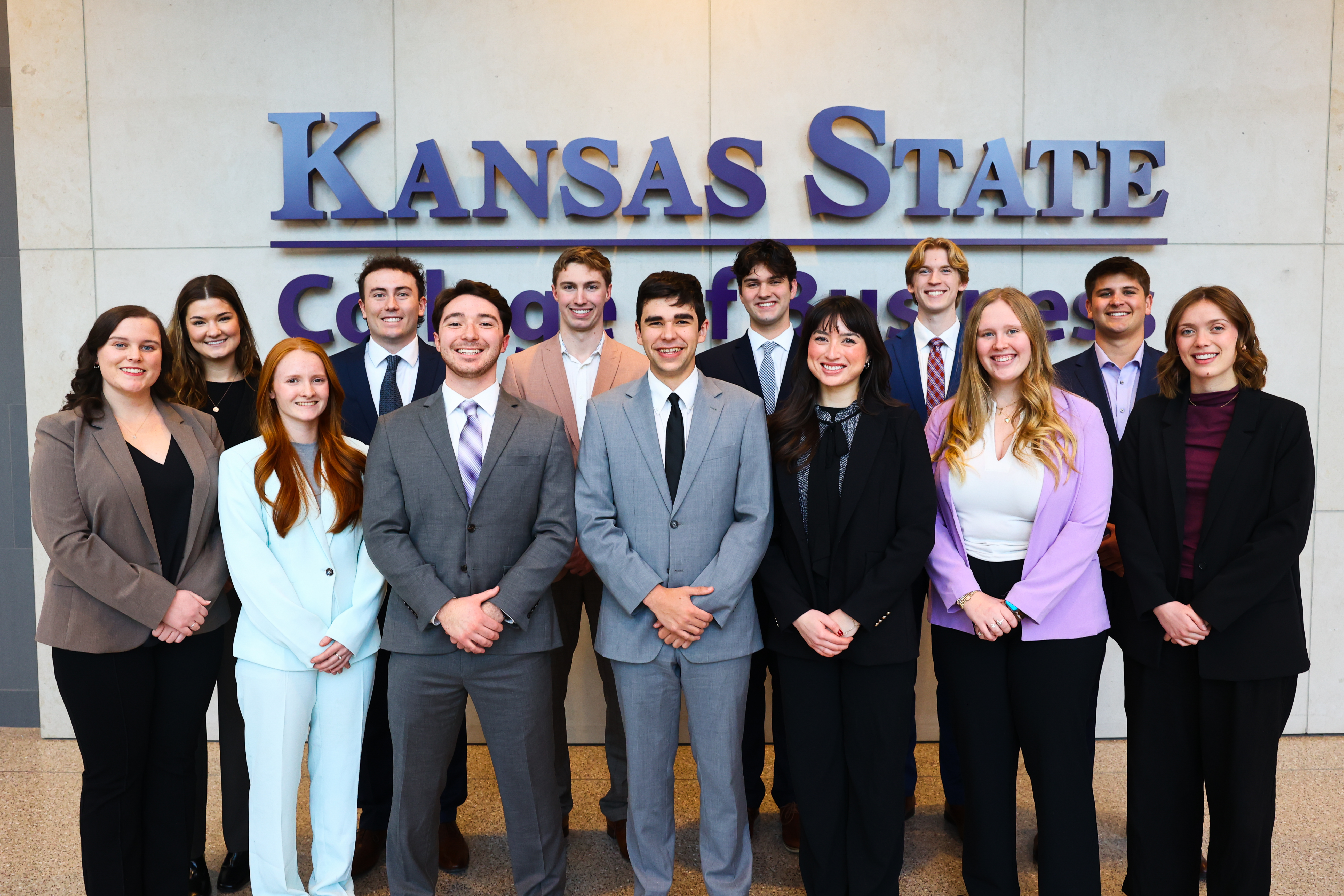 A group of 13 students in professional business attire stand in front of a Kansas State University wordmark.