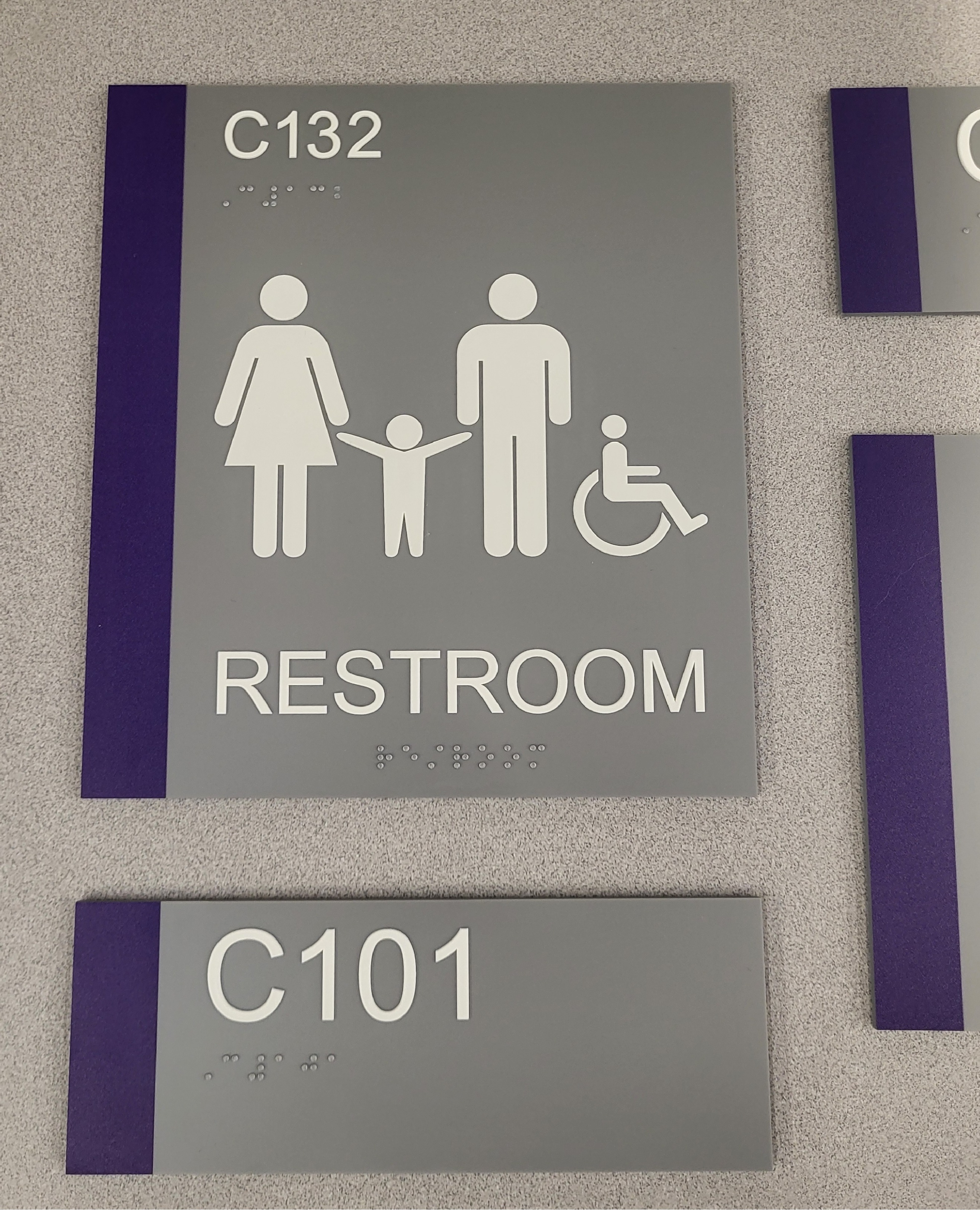 Luna GolTwo gray signs with purple bars on the left, the one on top a restroom sign showing a child between two people and a person in a wheelchair with Braille on the bottom and the one on the bottom a room number "C101" with Braille beneath.dendoodle Sougéy poses for a portrait.