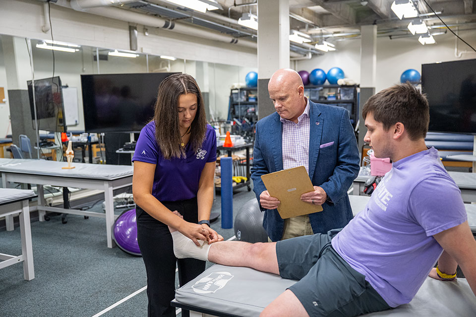 Student wearing a purple shirt wraps the ankle of another student wearing a purple shirt.  