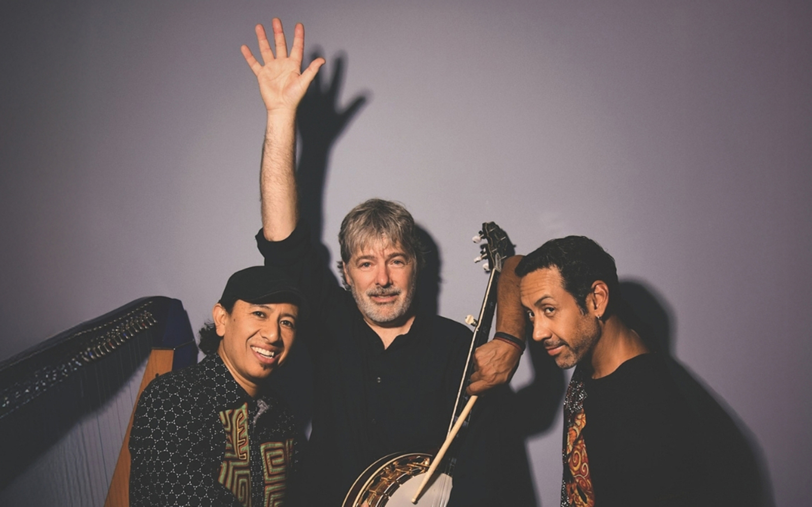 Three men in dark clothing are pictured against a light purple shadowy background, one with a banjo and one with a harp behind him.