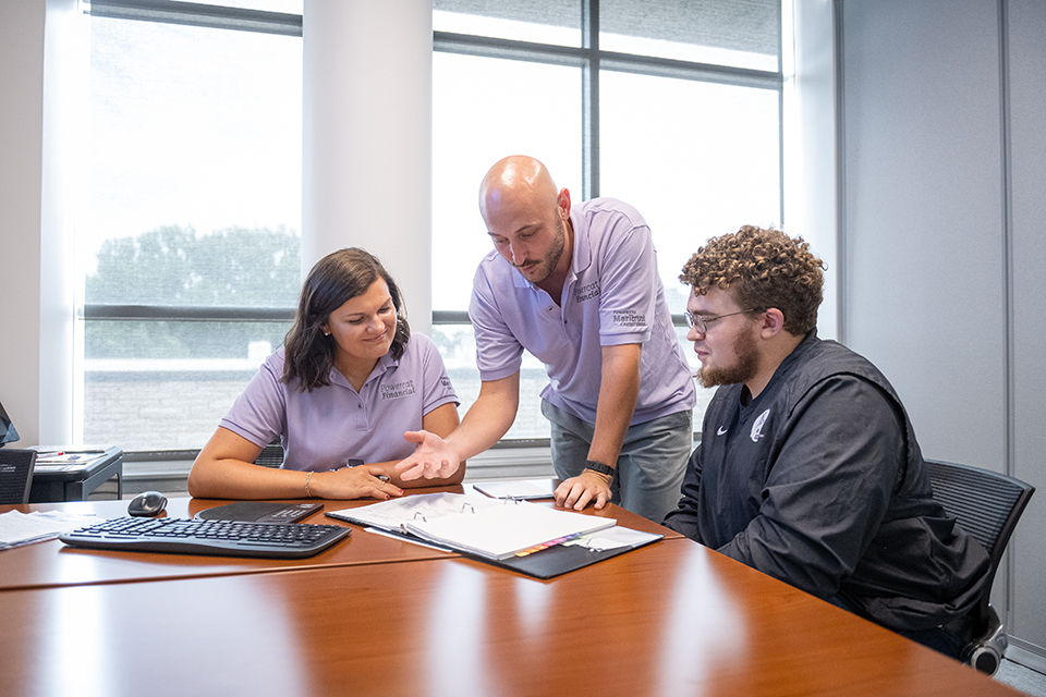 A financial educator in a lavender polo coaches students at a wooden table.