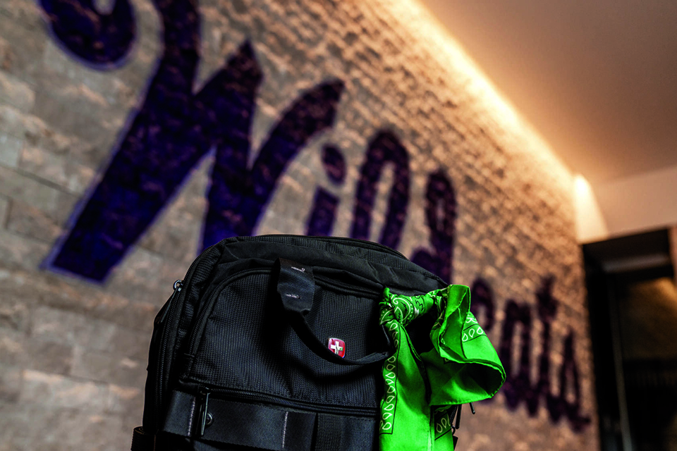 A black backpack with a green bandana tied to it sits in front of a white wall that has the word "Wildcats" written on it in stylized script.