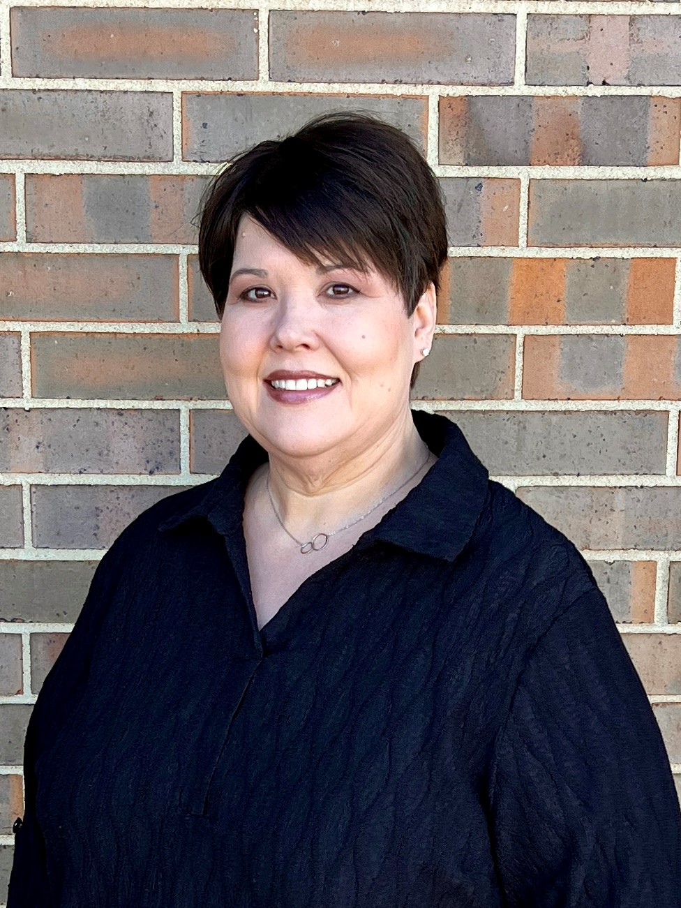 A woman with short, dark hair and a black shirt poses for a portrait in front of a brick wall.