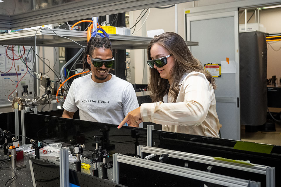 Students in light colored shirts wear eye protection and work in a physics lab. 