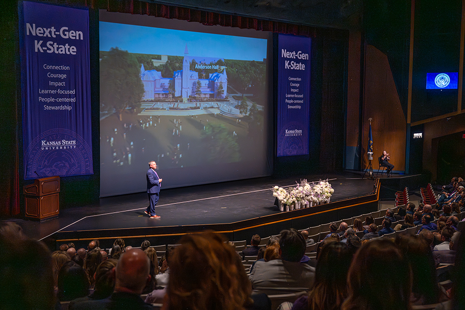 Looking down on an auditorium stage, a crowd listens to a presentation made by Kansas State University President Richard Linton. 