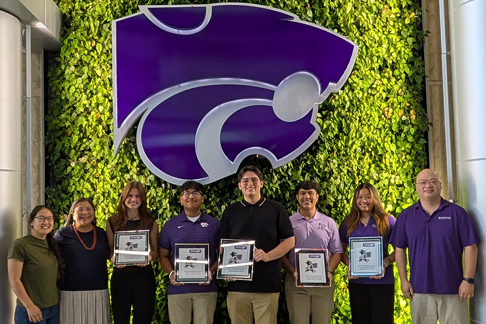 Seven people pose beneath the K-State Powercat logo and green garden wall, with several people holding award plaques.