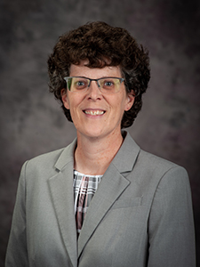 A woman with short, curly brown hair and glasses poses for a professional headshot in a gray blazer. 