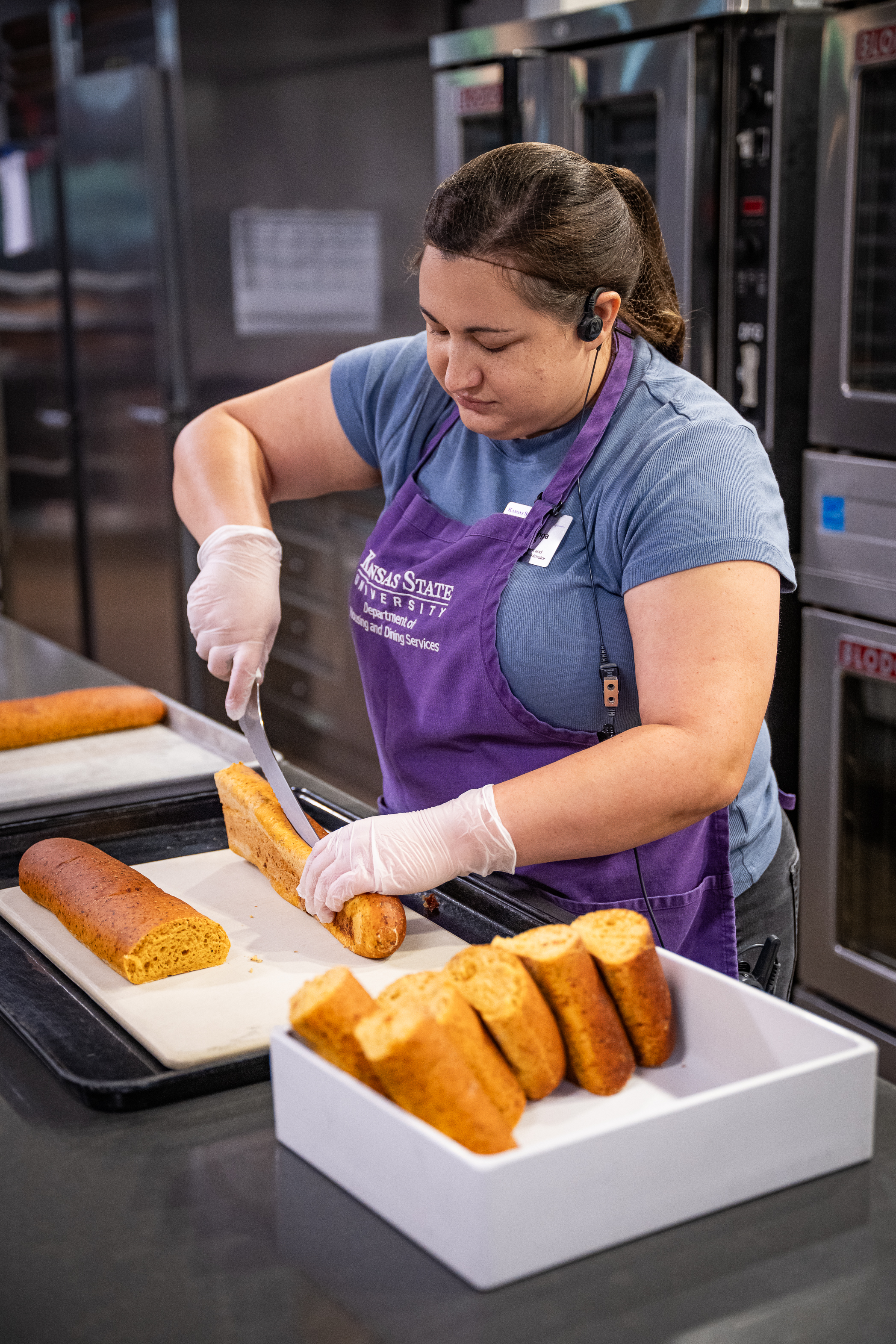 A student wearing a purple apron slices a loaf of bread lengthwise using a knife.