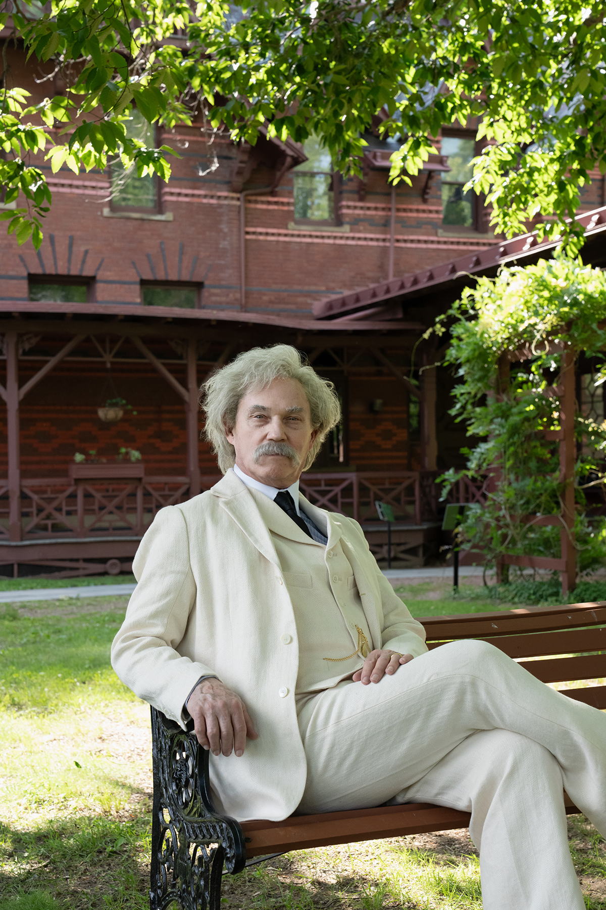 A man dressed as Mark Twain in a cream-colored suit and a navy tie sits on a bench oustide beneath a green trea in front of a red house.