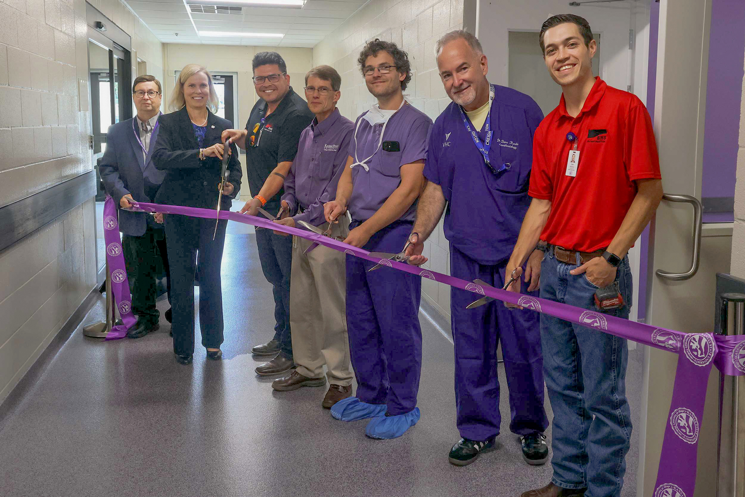 A group of seven people stand in a line behind a purple ribbon. Five people hold scissors to cut the ribbon.