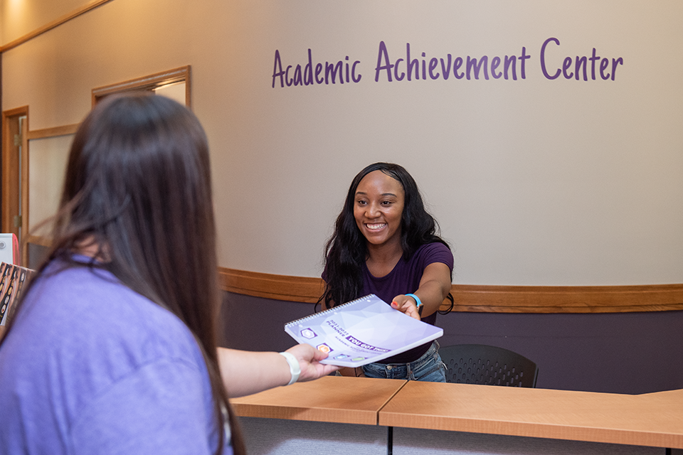 A college student reaches over a reception desk to hand a planner to the person working at the desk. The wall behind the desk has a sign that says Academic Achievement Center.