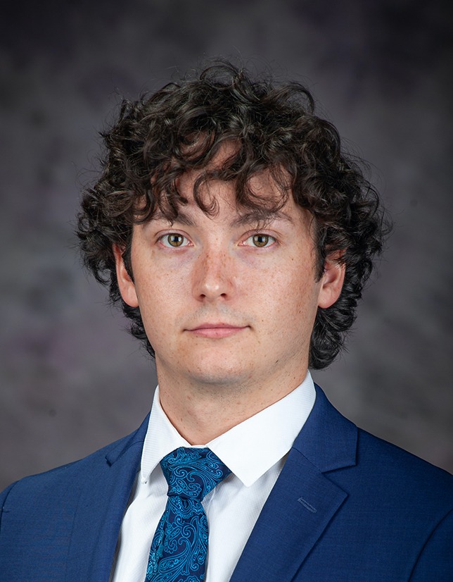 A young man with dark curly hair wears a blue suit with a blue patterned tie.