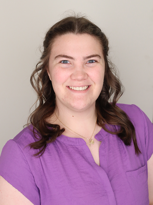 A young woman with long brown hair wears a lavender dress shirt against a gray background.