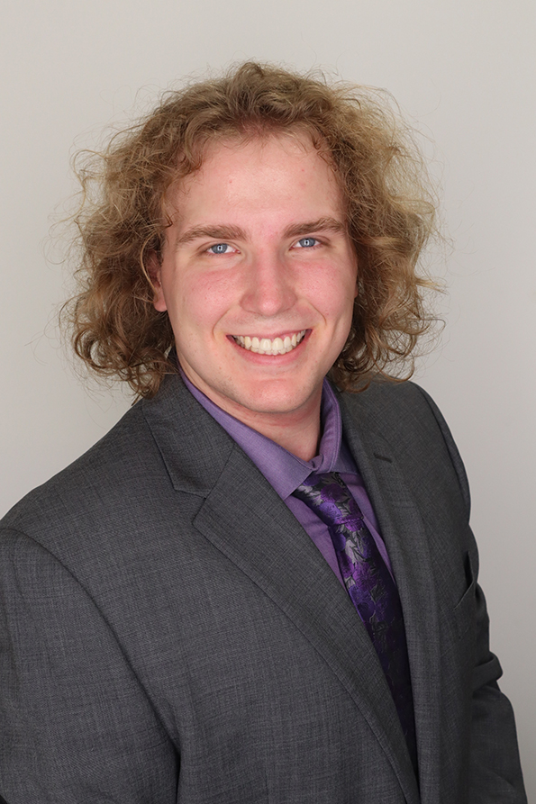 A young man with curly blonde/brown hair wears a gray suit with a purple shirt and tie.