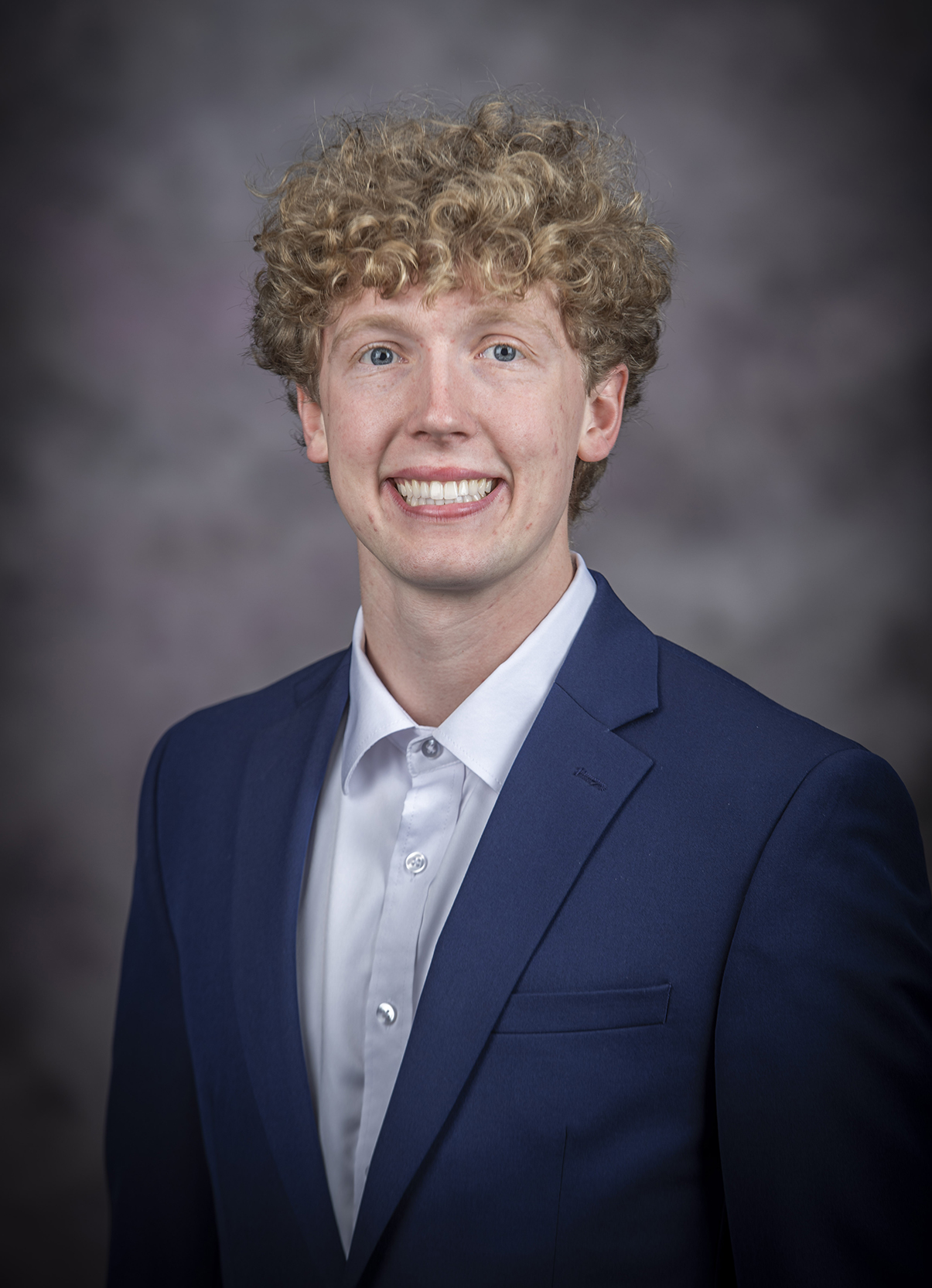 A young man with blonde curly hair wears a navy suit with a light blue shirt beneath.