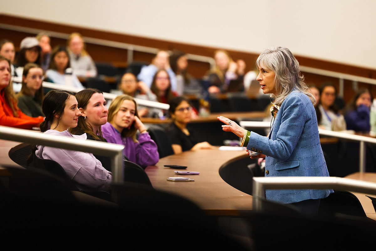 A woman with gray hear wearing a blue blazer speaks to a classroom of students.