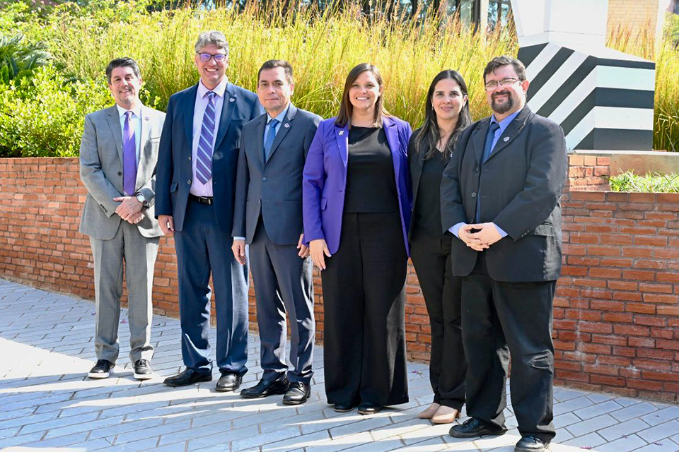 Six people in business professional attire stand outside on a sunny day for a group photo.