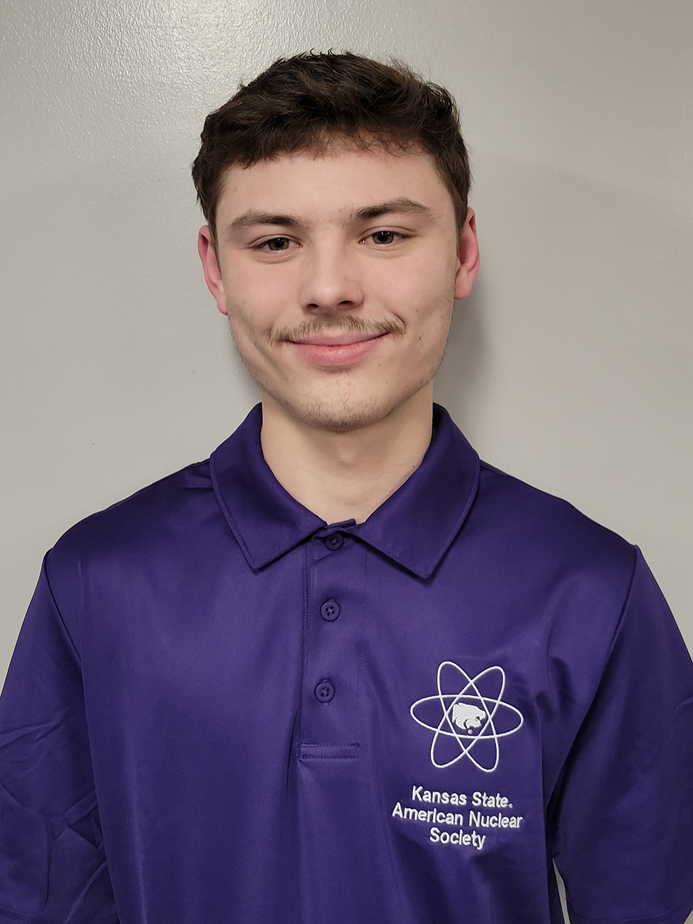 A young man with short, dark hair and a mustache wears a purple Kansas State American Nuclear Society polo.