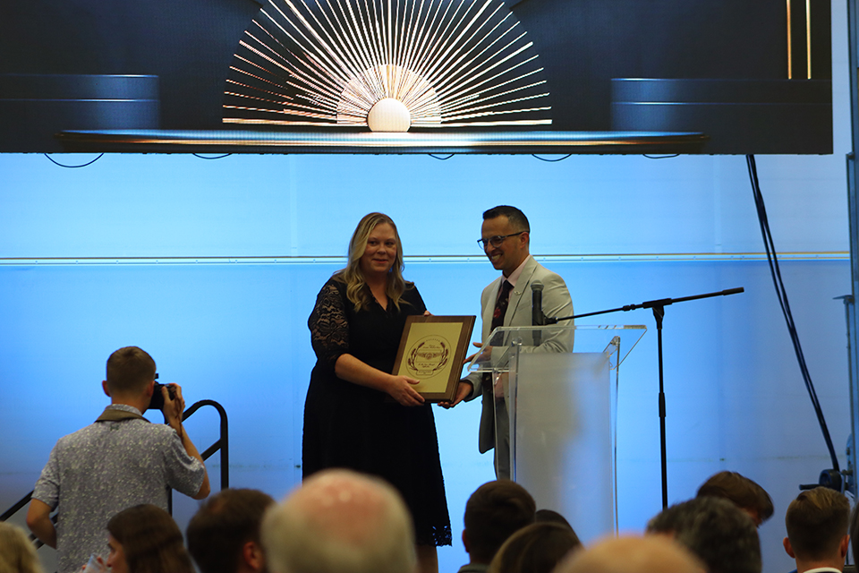 On a stage with a blue backdrop, a woman receives a plaque from a man while they pose for a photo taken by another photographer.