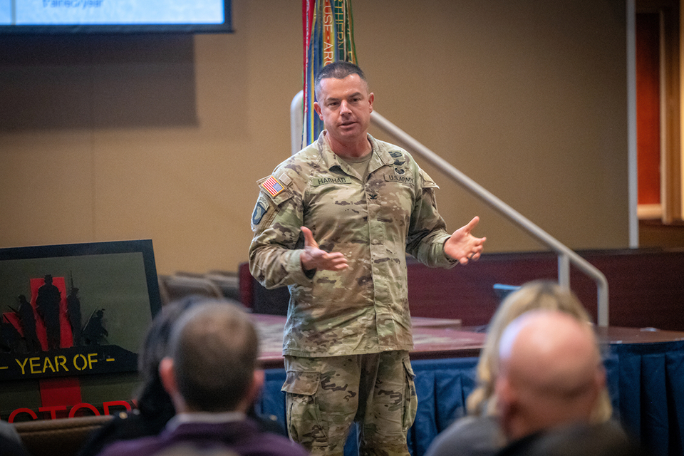 Col. Travis Habhab addresses a crowd in a meeting room. Behind him is a stage with a projector screen and various flags. 