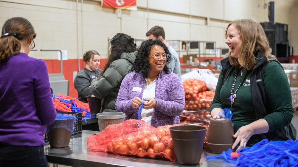 People are shown standing around metal tables that have onions on them in a community engagement project. Two women are featured, one in a purple sweater and one in a green sweater, laughing and working together.