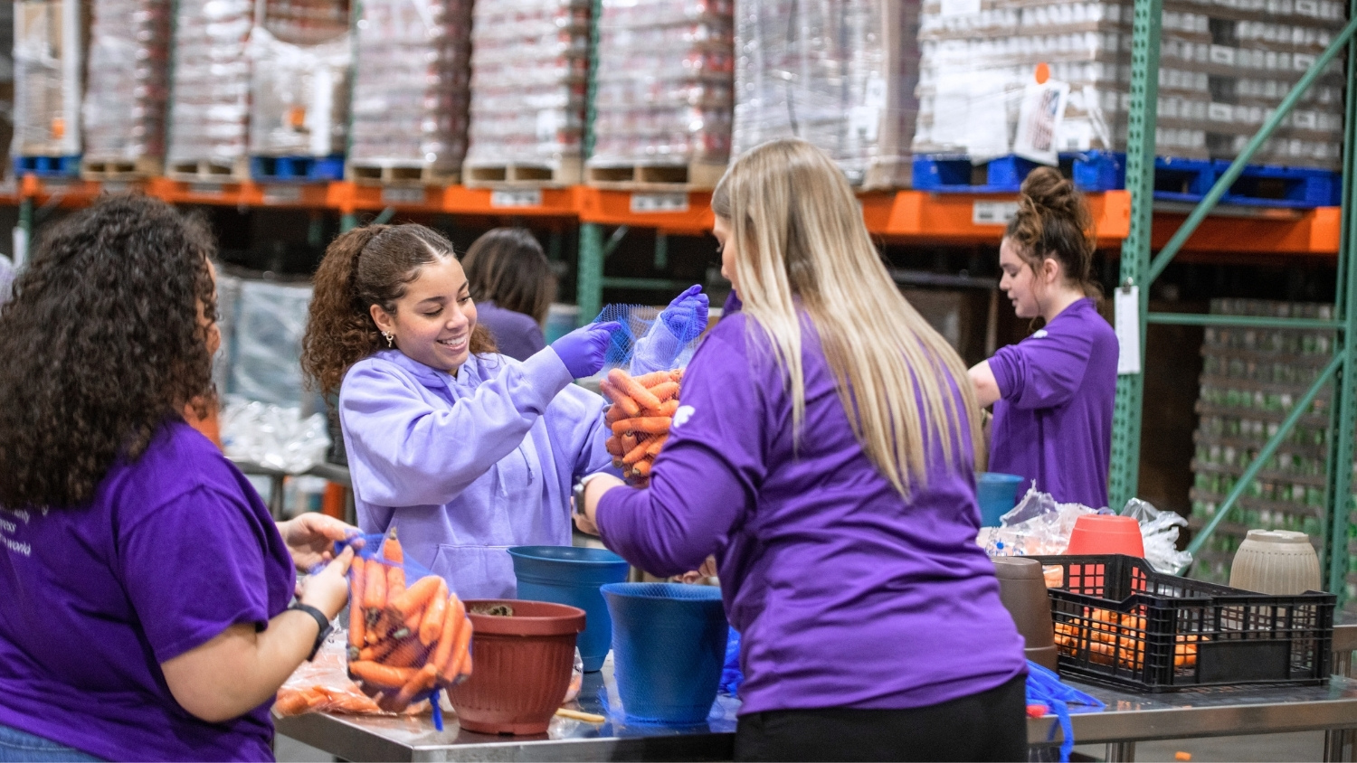 A young woman in a lavender hoodie bags carrots and holds them out to another young woman in a purple sweater who has her back to the camera.