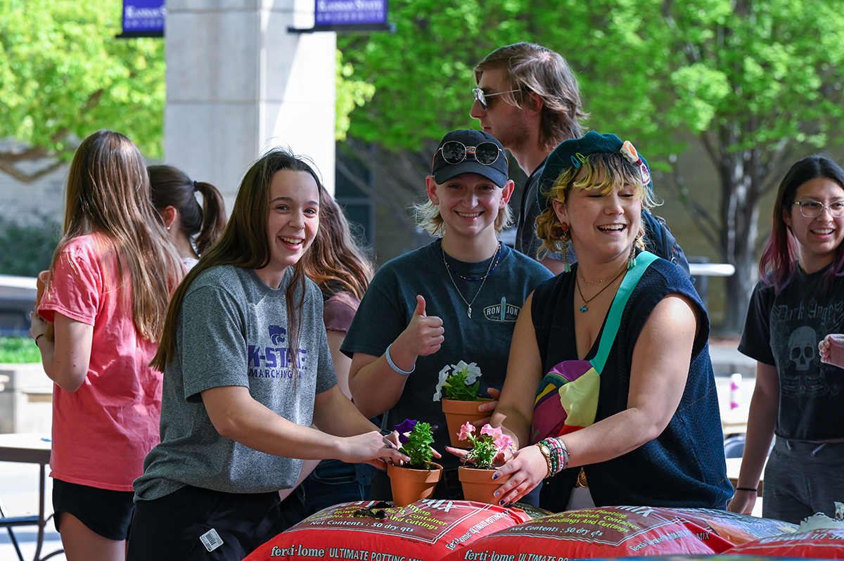 Three students are pictured standing behind bags of soil, each student holding newly potted flowers. On the left, a student with long brown hair wearing a gray T-shirt holds a purple flower; in the middle, a student with short blonde hair wearing a hat and sunglasses gives a thumbs up while holding white flowers; at right, a student with blonde and yellow-tipped hair wearing a beret and barrettes smiles to the side and holds pink flowers. Other students mill about in the background.